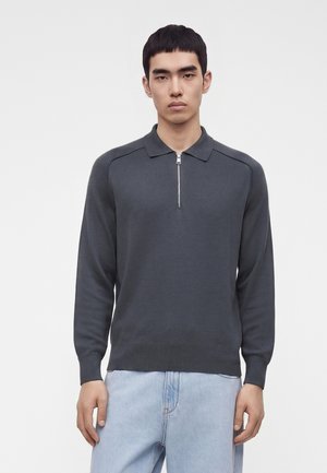 Jeune homme portant un pull polo à manches longues gris foncé avec une fermeture éclair à l'avant et un jean bleu clair, debout devant un fond uni.