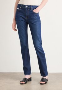 Jeans de denim azul-escuro com um corte slim, apresentando fecho de botão, bolsos frontais e detalhes de costura subtis. Combinados com sandálias pretas de salto bloco.