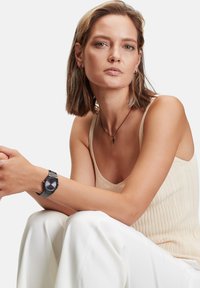 Schwarze Uhr mit rundem Ziffernblatt und silbernen Akzenten, am Handgelenk getragen. Modell in einem beigefarbenen gerippten Oberteil und weißen Hosen, sitzend in entspannter Pose.