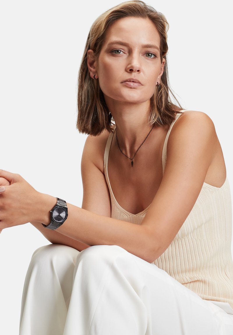 Schwarze Uhr mit rundem Ziffernblatt und silbernen Akzenten, am Handgelenk getragen. Modell in einem beigefarbenen gerippten Oberteil und weißen Hosen, sitzend in entspannter Pose.