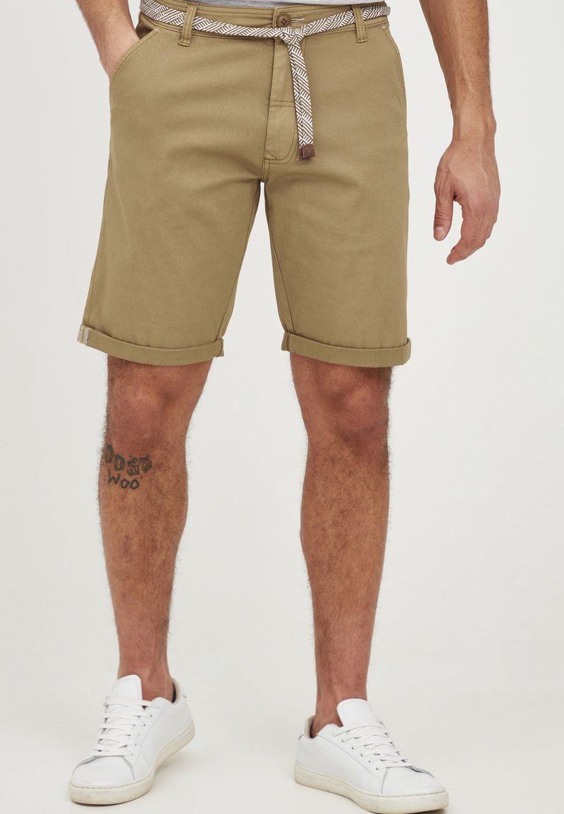 Bărbat îmbrăcat cu pantaloni scurți khaki suflecați, cu o curea din material căptușit, adidași albi și un tatuaj pe gambă stângă.