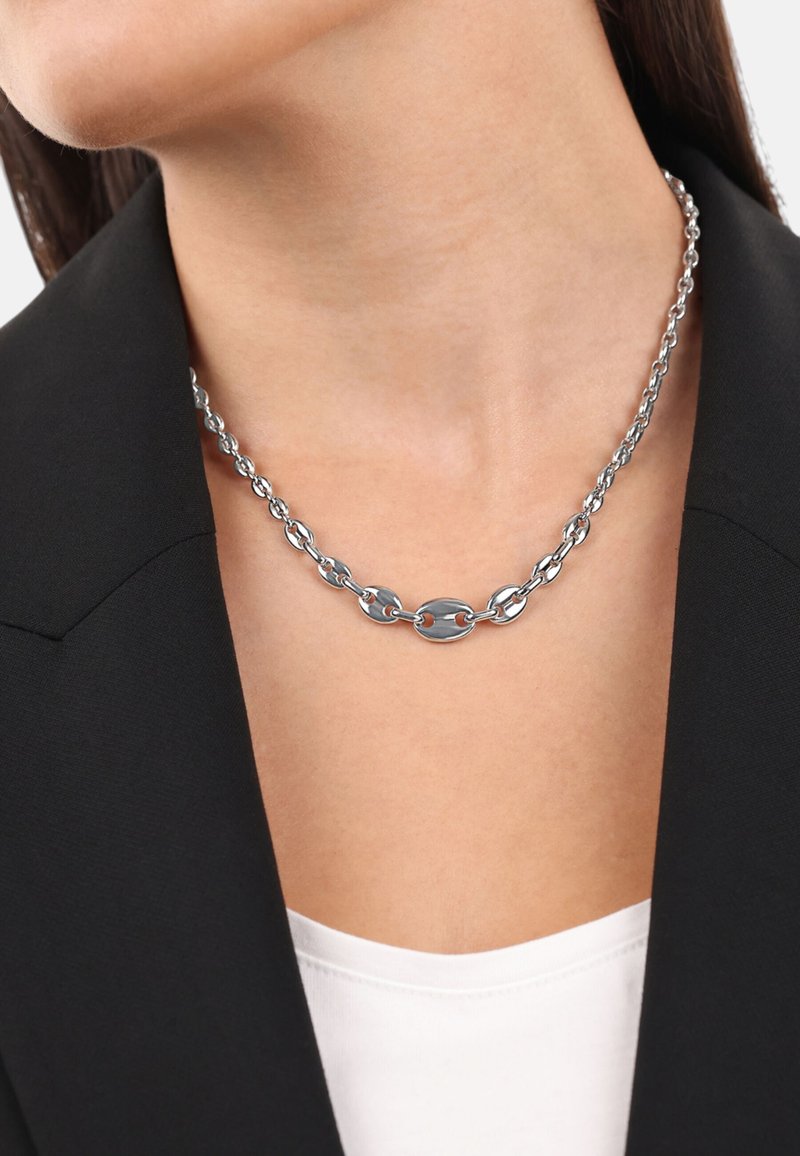 Collana in argento caratterizzata da anelli ovali intrecciati, finitura lucida e una chiusura decorativa centrale. Indossata sopra una maglietta bianca e un blazer nero.
