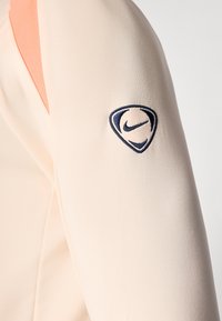 Beige and peach long-sleeve garment sleeve with a dark blue embroidered Nike swoosh inside a shield-shaped outline.