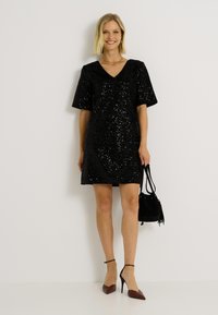 Robe noire à sequins avec un décolleté en V et des manches courtes. Associée à des escarpins à bout pointu et un petit sac seau noir. Texture lisse et détail brillant.