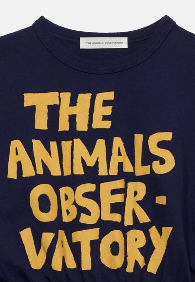 Μπλε ναυτικό πουκάμισο με μεγάλο, άνισο κίτρινο κείμενο που γράφει "THE ANIMALS OBSERVATORY" και μια μικρή λευκή ετικέτα με το όνομα της μάρκας μέσα στον γιακά.
