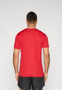 Punane lühikeste varrukatega sportlik T-särk, sileda tekstuuriga, ümmarguse kaelakaarega ja vormi järgiva lõikega. Kandjana mustad lühikesed püksid, vaadatuna tagantpoolt.