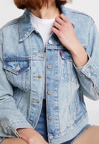 Světle modrá denimová bunda s klasickým designem, která má dvě náprsní kapsy, stříbrné knoflíky a opotřebované detaily na látce.