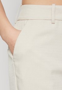 Pantalons texturés beiges avec une large ceinture, dotés d'une poche latérale et d'un cordon de serrage pour l'ajustement. Tissu doux avec une apparence tissée.
