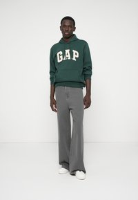 Zielona bluza z kapturem z logo "GAP" w kolorze kremowym, zestawiona z szarymi spodniami w szerokim kroju. Model nosi białe sneakersy, stojąc na jednolitym tle.