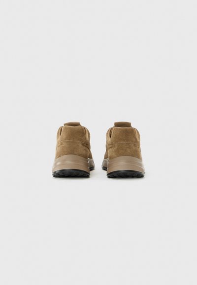O pereche de adidași maro din suede văzuți din spate pe un fundal alb, arătând tălpi groase cu profil negru.