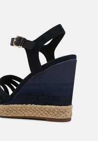 Sandale compensée bleu marine avec semelle en jute tressée, bride noire à la cheville avec boucle dorée et signature embossée sur le talon.