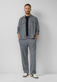 Mężczyzna stoi przy jasnoszarej ścianie, ubrany w szary taliowany żakiet, pasujące spodnie, czarną koszulę, białe sneakersy, ręce w kieszeniach, neutralny wyraz twarzy.