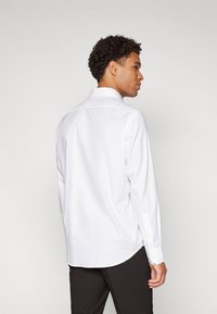 Chemise blanche à manches longues avec un col classique, texture lisse et poignets boutonnés ; la vue arrière montre un ourlet courbé et une coupe ajustée.