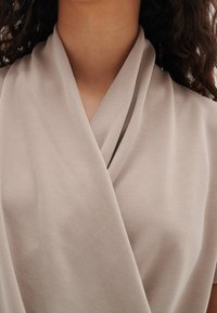 Femme portant un vêtement en tissu drapé beige, mettant en avant l'encolure et la partie supérieure de la poitrine avec des mèches de cheveux bouclés visibles.