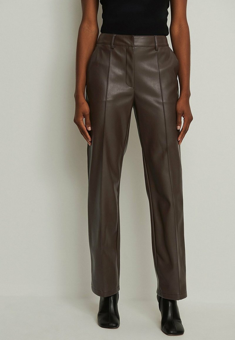 Bruna byxor i konstläder med rak passform, slät yta, framfickor och diskreta sömmar; kombinerat med svarta ankelskor.