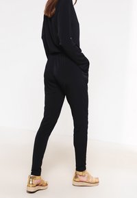 Černý overal s volným střihem, dlouhými rukávy a bočními kapsami. Nosený se zlatými sandály na páskovém opatku a tkanou platformou.