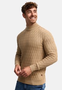 Pull en maille beige avec un col montant, motif texturé, poignets et ourlet côtelés, arborant un petit patch de marque sur le côté.