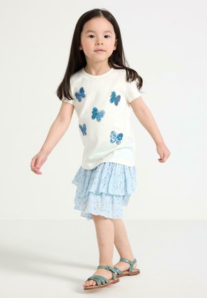 Ung pige står, iført en hvid skjorte med blå paillet sommerfugle, et lyseblåt lagdelt nederdel og teal sandaler mod en ensfarvet baggrund.