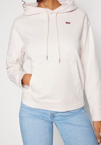 Hoodie rose clair avec une poche frontale, capuche à cordon de serrage et un petit logo rouge. Fabriqué en tissu doux, il présente des manches longues et des poignets côtelés.