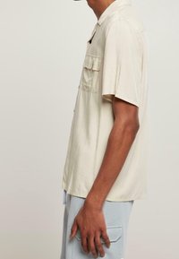 Homme portant une chemise beige à manches courtes avec des poches poitrine et un pantalon bleu clair avec des poches sur le côté, vu de profil.