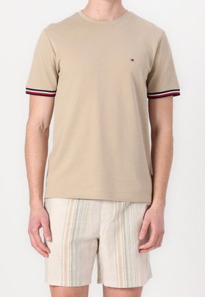 Mężczyzna w beżowej koszulce z krótkim rękawem z czerwonym, białym i czarnym wykończeniem na rękawach oraz jasnych szortach w paski.