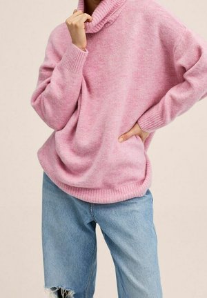 Osoba nosící volný růžový rolák a světle modré roztrhané džíny, jednou rukou si sahá na bradu, druhou má na boku.