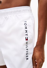 Fehér úszónadrág, rugalmas derékpánttal, kék és piros hímezett "TOMMY HILFIGER" logóval a bal oldalon, sima szövettextúrával.