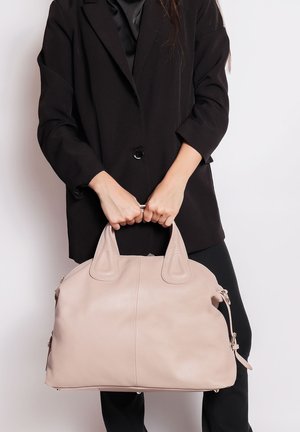 Persona che indossa un blazer nero e pantaloni, tiene una grande borsa in pelle beige chiaro con due manici davanti a uno sfondo bianco.