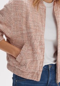 Jasnoróżowa bomberka z teksturowanej tkaniny, elastycznymi mankietami i zamkiem z przodu. Posiada boczne kieszenie i luźny krój.