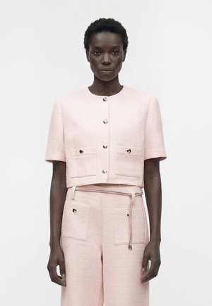 Žena koja nosi blijedoružičastu teksturiranu kratku jaknu na kopčanje i odgovarajuće hlače visokog struka s prednjim džepovima i platnenim remenom.