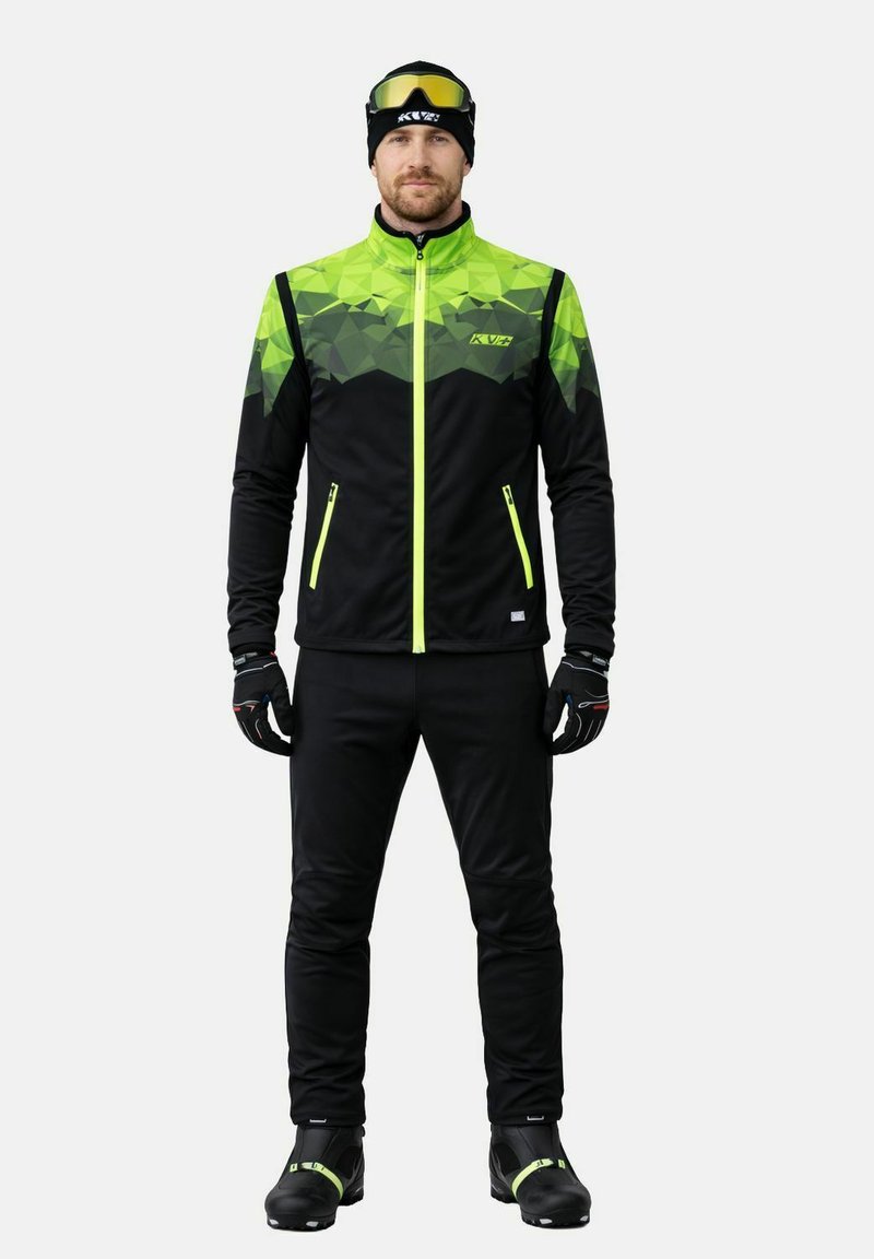 Homme portant une veste de sport noire et vert néon, pantalon noir, gants, bonnet, chaussures de ski et lunettes de ski à verres teintés jaunes sur la tête.