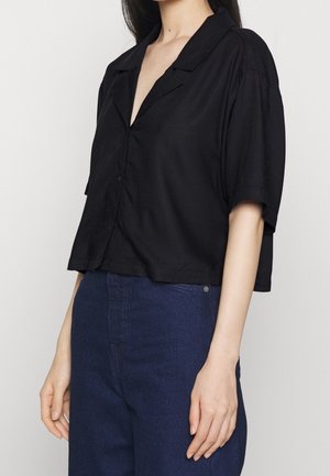 Kobieta ubrana w luźną czarną koszulę z krótkim rękawem i kołnierzykiem, wsuniętą w spodnie z wysokim stanem w ciemnoniebieskim kolorze, na jednolitym tle.