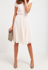 Une robe beige, longueur genou, avec des manches courtes, des plis et une taille cintrée. Associée à des escarpins gris et tenant une pochette argentée.