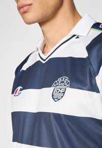 Mand iført en navy og hvid stribet sportsjersey med en hvid krave og logoer på brystet, som vender let mod venstre.