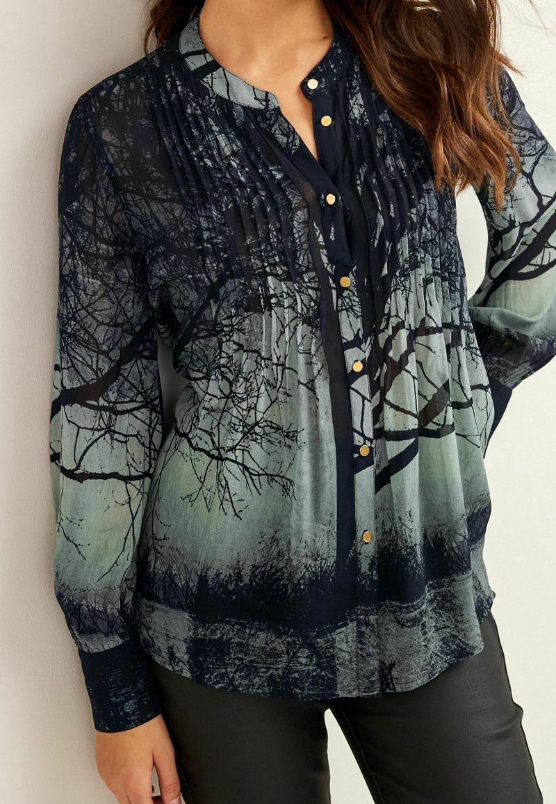 Blouse à manches longues avec boutons, motif sombre de branches d'arbre sur un fond dégradé gris et vert, portée avec un pantalon noir.