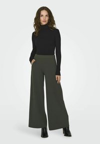Top a collo col ribbed nero abbinato a pantaloni larghi a vita alta color verde oliva con tasche laterali. La modella indossa stivali neri con tacco.