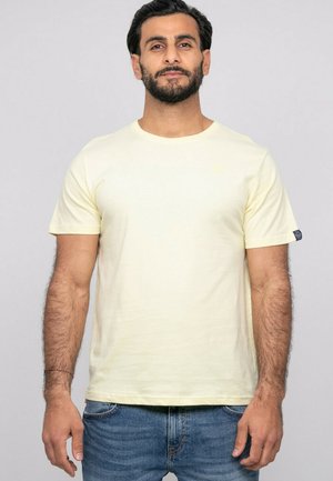 Homme aux cheveux foncés et à la barbe, portant un t-shirt jaune clair uni à manches courtes et un jean bleu, debout devant un fond clair uni.