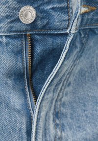 Gros plan d'un jean en denim bleu avec une fermeture éclair en laiton partiellement ouverte et un bouton argenté portant l'inscription "MNG DENIM".