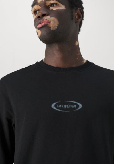 Camiseta de manga larga negra con cuello redondo. Presenta un sutil logo azul en el pecho y una textura suave, similar al algodón.