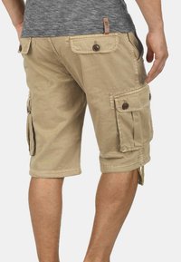 Beige Cargo-Shorts mit mehreren knopfverschlossenen Taschen, getragen von einer Person, die steht und die Rück- und Seitenansicht zeigt.