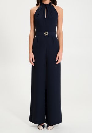 Woman wearing a sleeveless navy halter jumpsuit with wide legs and a decorative belt buckle, paired with two-tone pointed shoes.