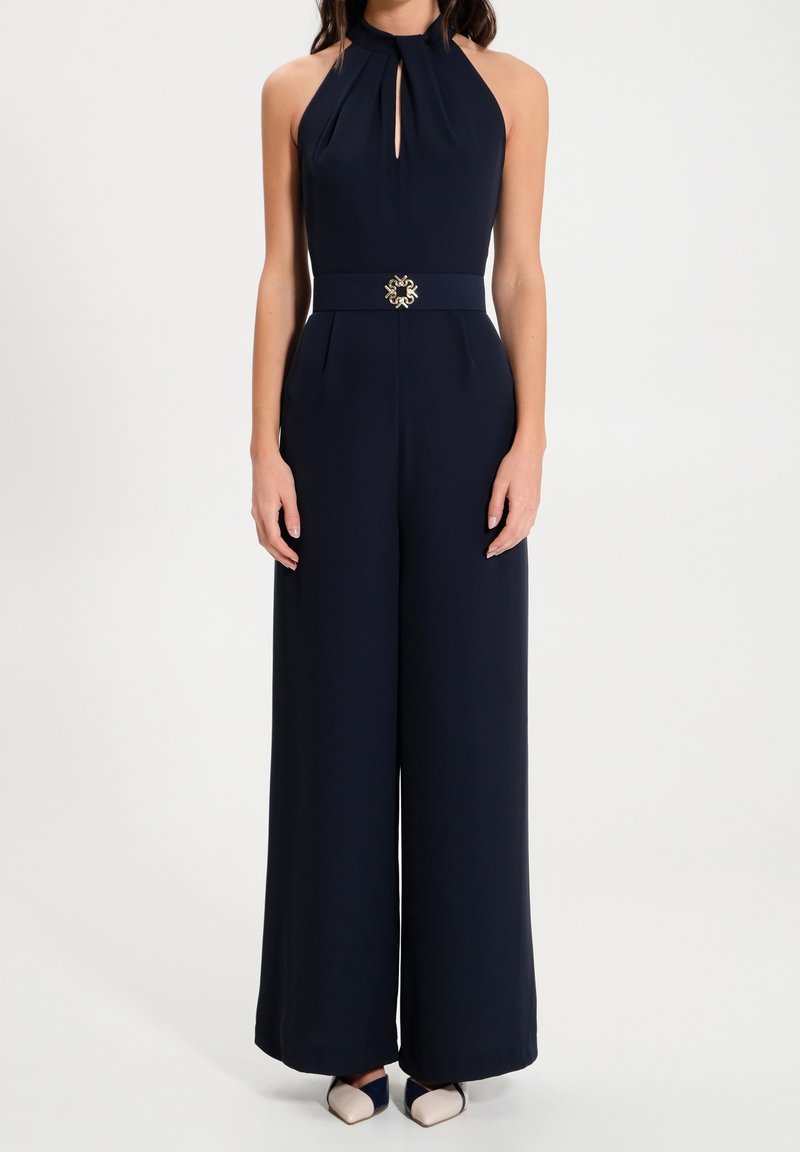 Femme portant une combinaison sans manches bleu marine à col halter avec jambes larges et une boucle de ceinture décorative, associée à des chaussures pointues bicolores.