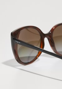 Ochelari de soare cu gradient maro, cu rame supradimensionate și rotunde. Prezintă un braț din metal negru contrastant cu brandul "Vogue".