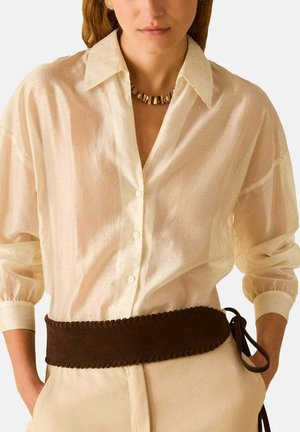 Vrouw draagt een crème-textuur overhemd met knopen, een kralenketting en een brede donkerbruine riem die aan de zijkant is gestrikt, met handen in de zakken.