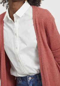 Rosa kardiagan över en vit skjorta med en texturerad knappslå, i kombination med blå jeans. Tyget ser mjukt och mysigt ut.