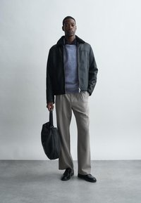 Férfi fekete bőrkabátot, kék pulóvert, bézs nadrágot és fekete cipőt visel, miközben egy fekete bőr táskát tart egy sima világos háttér előtt.