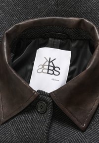 Manteau à motif chevrons gris foncé avec un col en cuir marron foncé et une étiquette blanche visible à l'intérieur indiquant "Karen by Simonsen."