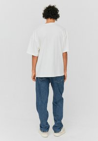 T-shirt blanche surdimensionnée à manches courtes, présentée de dos. Portée avec un jean bleu et des chaussures claires. Texture en coton lisse.