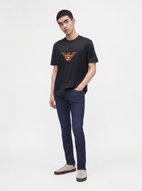 Млад мъж, носещ черна тениска Emporio Armani, тъмносини дънки и бежови маратонки, стои с една ръка в джоба си на бял фон.