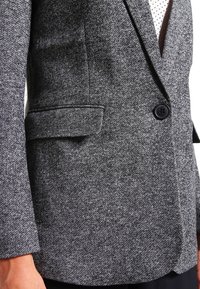Blazer gris sur mesure en tissu texturé, avec une fermeture à un bouton et une poche sur le côté gauche.
